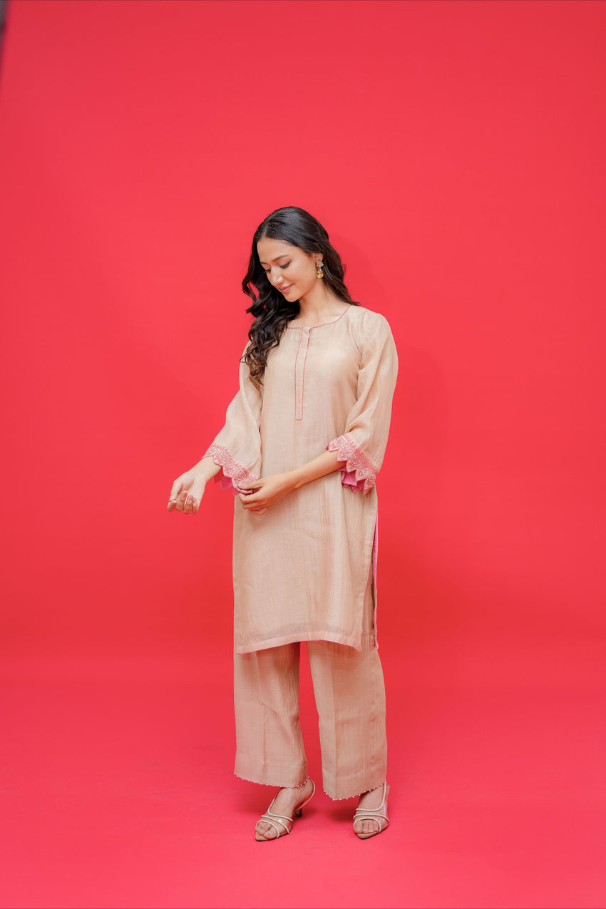 Female model wearing a light beige ethnic kurta and palazzo set with delicate embroidery and pink lace accents on the sleeves, standing against a bright red studio backdrop.