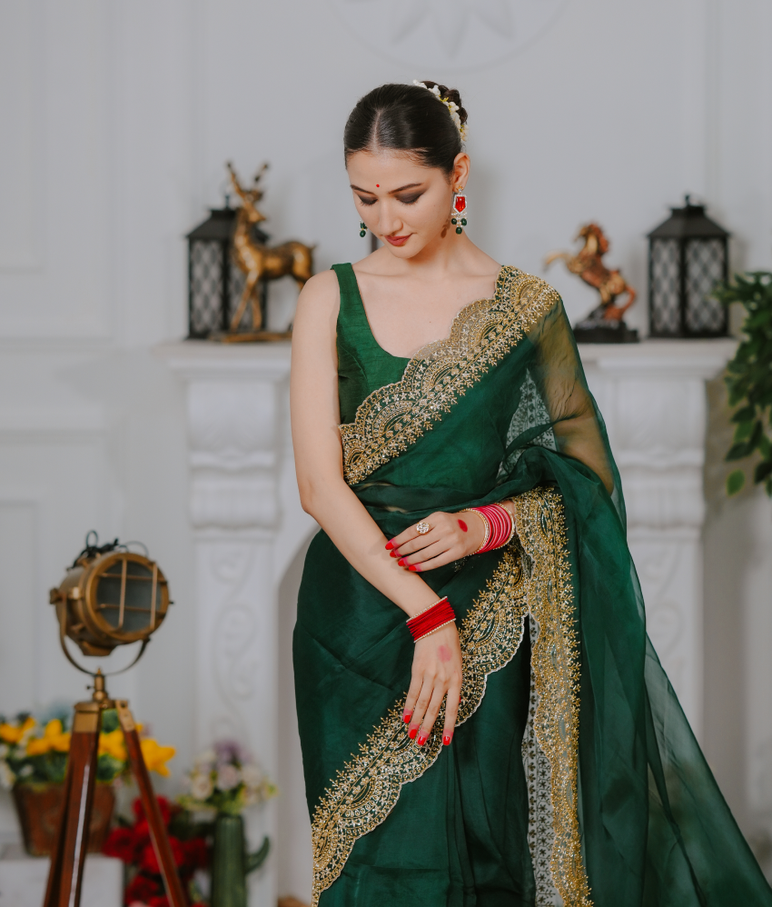 Woman in a green saree with gold border standing in a decorated room.